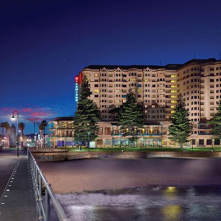 Stamford Grand Adelaide Hotel Гленелг Екстериор снимка The photo shows a waterfront scene at dusk, featuring a large building lit up against a darkening sky. The architecture appears modern and prominent, with several windows illuminated. In the foreground, there is a walkway or pier with fencing along t