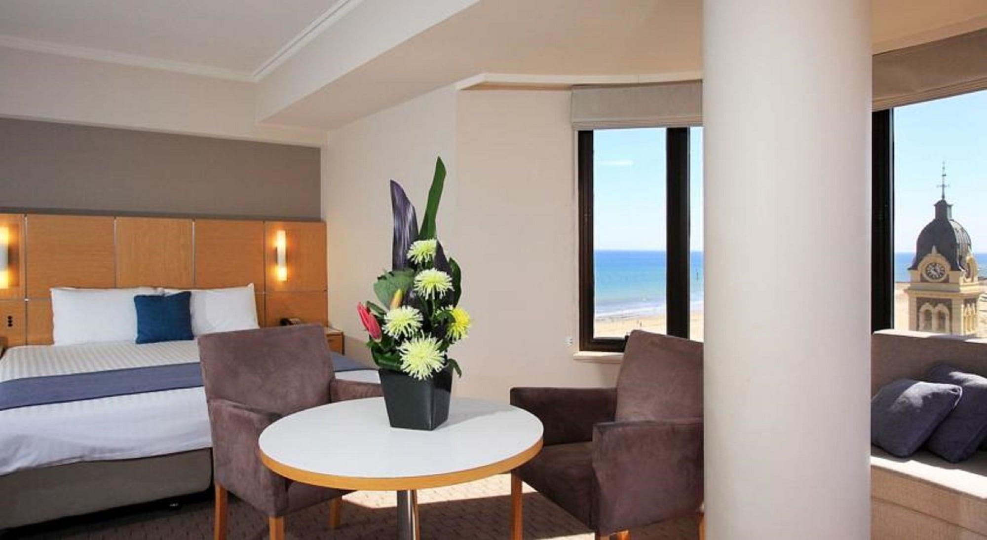 Stamford Grand Adelaide Hotel Гленелг Екстериор снимка The photo shows a cozy hotel room with a comfortable seating arrangement. There are two brown chairs positioned around a round table, which features a decorative flower arrangement in a black vase. The background includes a window that offers a view 