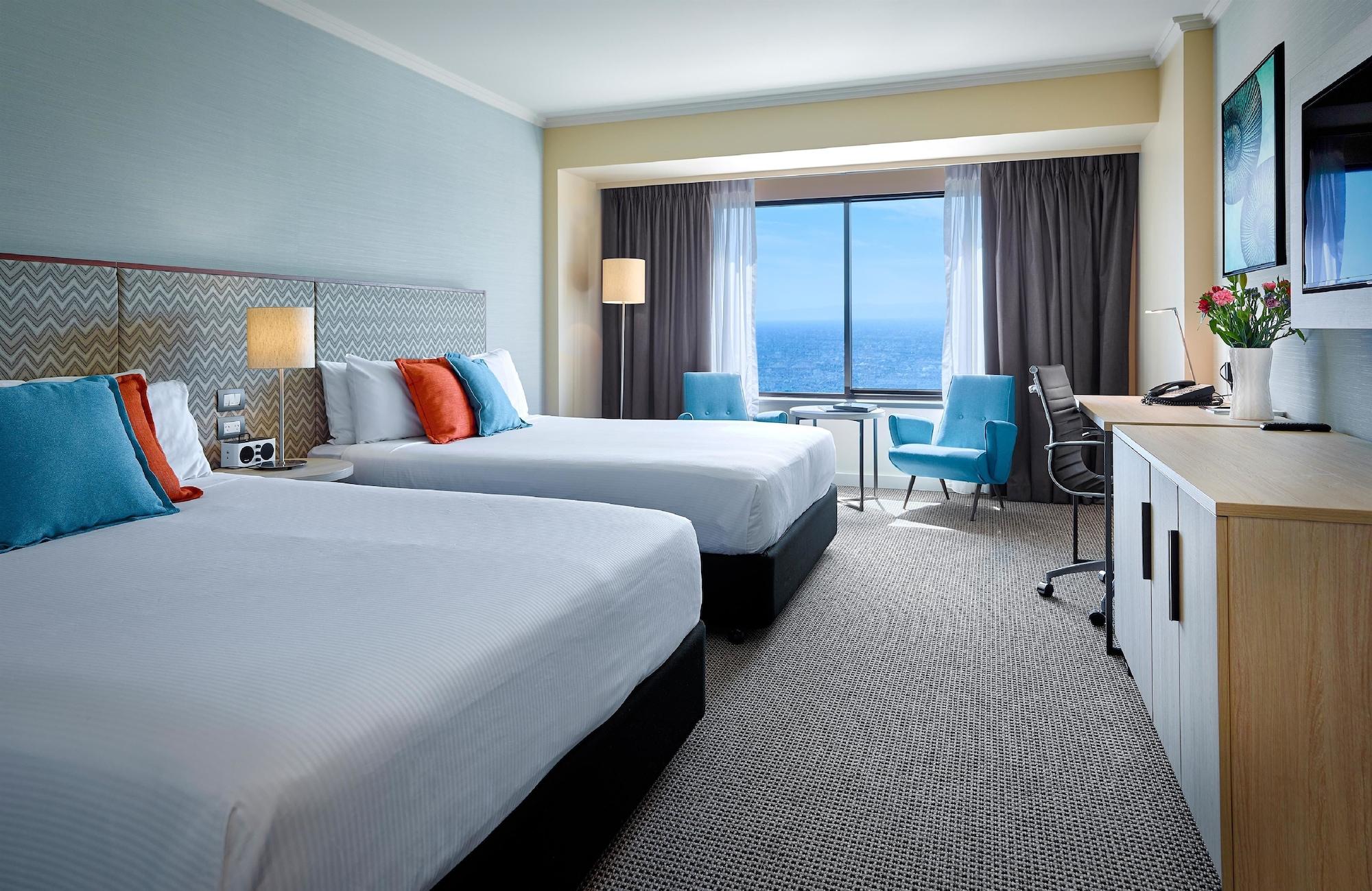 Stamford Grand Adelaide Hotel Гленелг Екстериор снимка The photo shows a well-decorated hotel room featuring two queen-sized beds with white linens and colorful accent pillows. There is a large window with curtains that offers a view of the ocean. A small seating area with a blue chair and a matching des