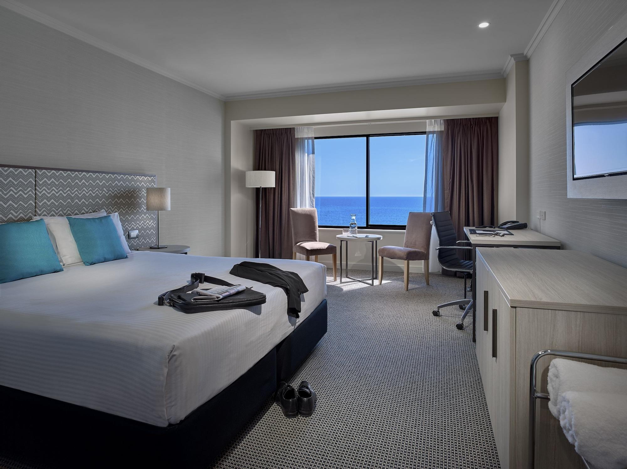 Stamford Grand Adelaide Hotel Гленелг Екстериор снимка The photo shows a modern hotel room featuring a queen-sized bed with white linens and two aqua blue decorative pillows. There are bedside lamps and a large window with a view of the ocean. The room also has a small seating area with two chairs and a 