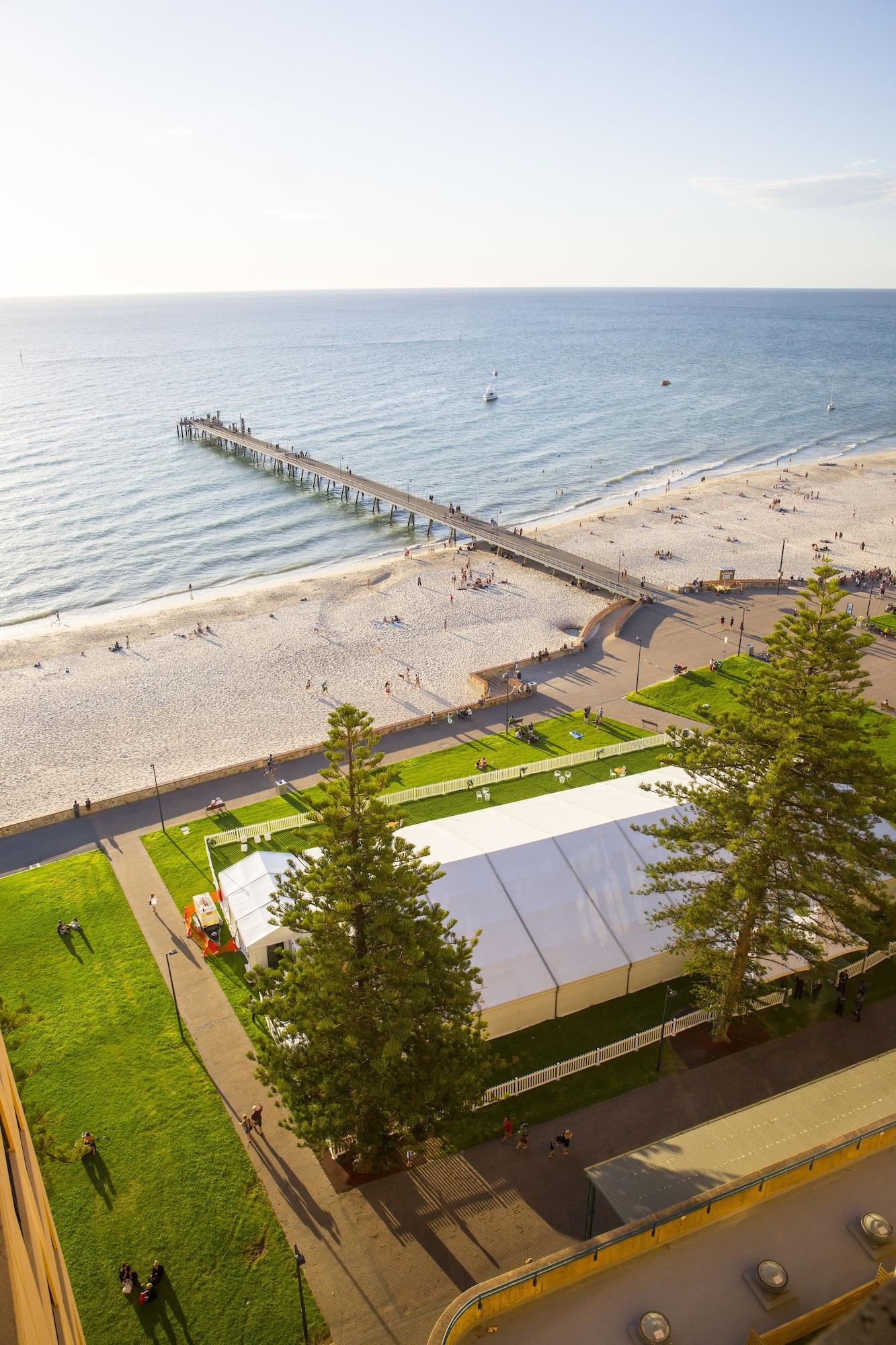 Stamford Grand Adelaide Hotel Гленелг Екстериор снимка The photo shows a scenic view of a beach with a long pier extending into the water. The beach is sandy, and there are people enjoying the sun and walking along the shore. In the foreground, there is a large white tent set up on a grassy area, likely 