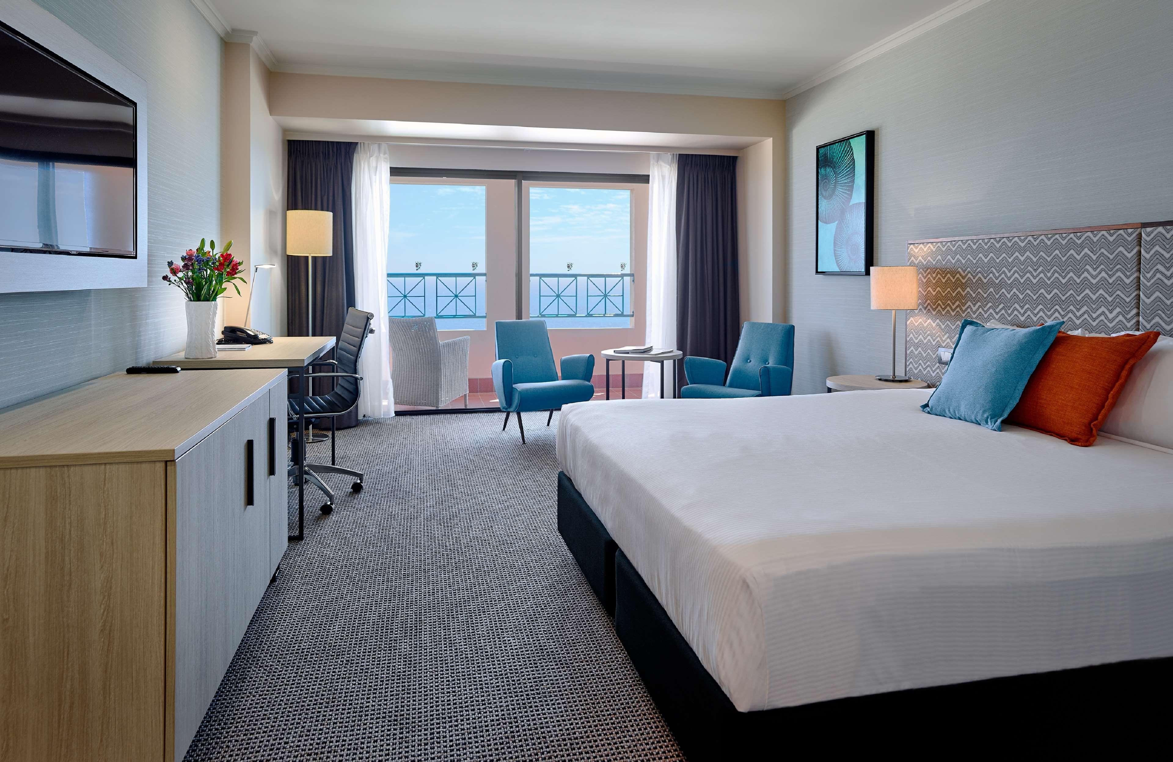 Stamford Grand Adelaide Hotel Гленелг Екстериор снимка The photo depicts a modern hotel room. The room features a large bed with a white comforter, and there are decorative pillows in orange and blue. There is a flat-screen TV mounted on the wall, and a desk with an office chair is positioned near the wi