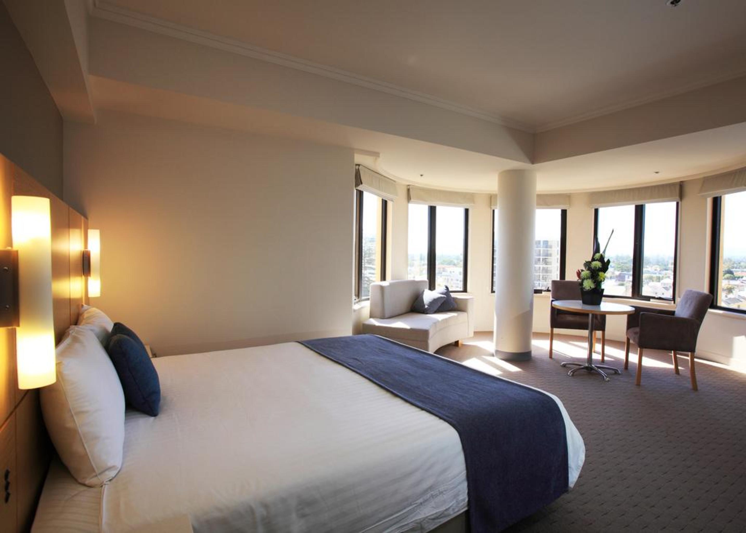 Stamford Grand Adelaide Hotel Гленелг Екстериор снимка The photo shows a hotel room with a modern and spacious design. There is a large bed with white linens and a dark blue throw blanket. The room features multiple windows that allow plenty of natural light, and there is a small seating area near the wi