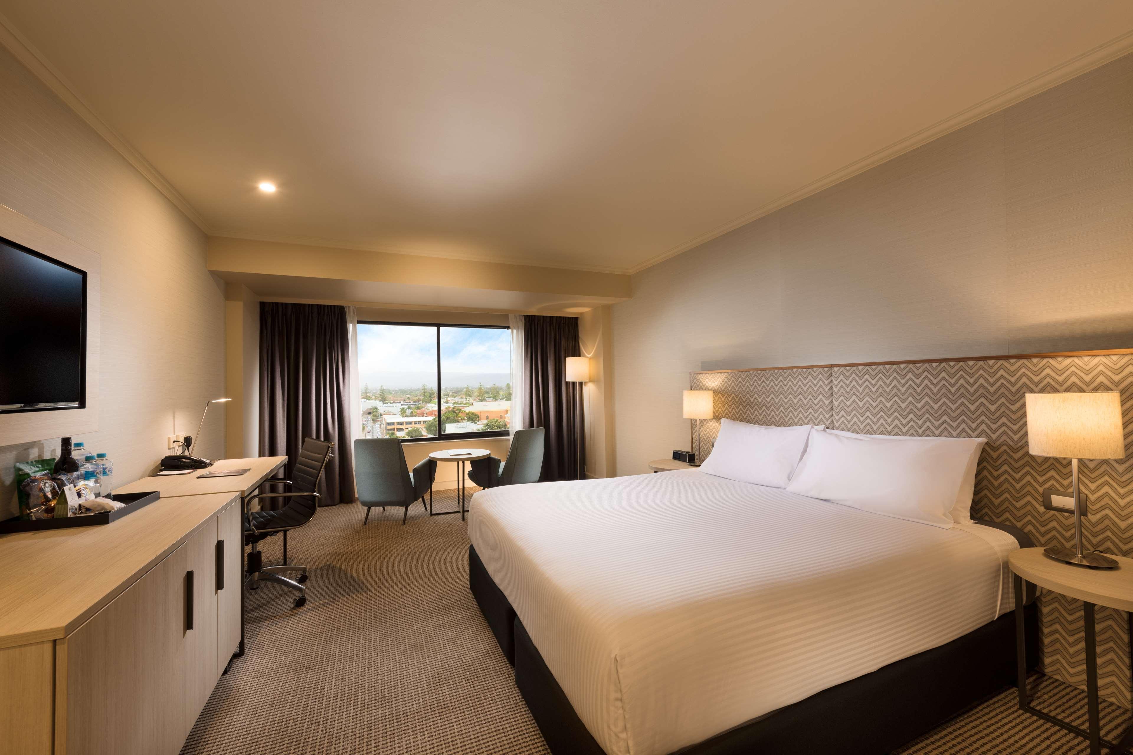 Stamford Grand Adelaide Hotel Гленелг Екстериор снимка The photo shows a modern hotel room. It features a large bed with white linens, a desk with a chair, and a flat-screen TV mounted on the wall. There are two armchairs and a small table near a large window that lets in natural light, providing a view 
