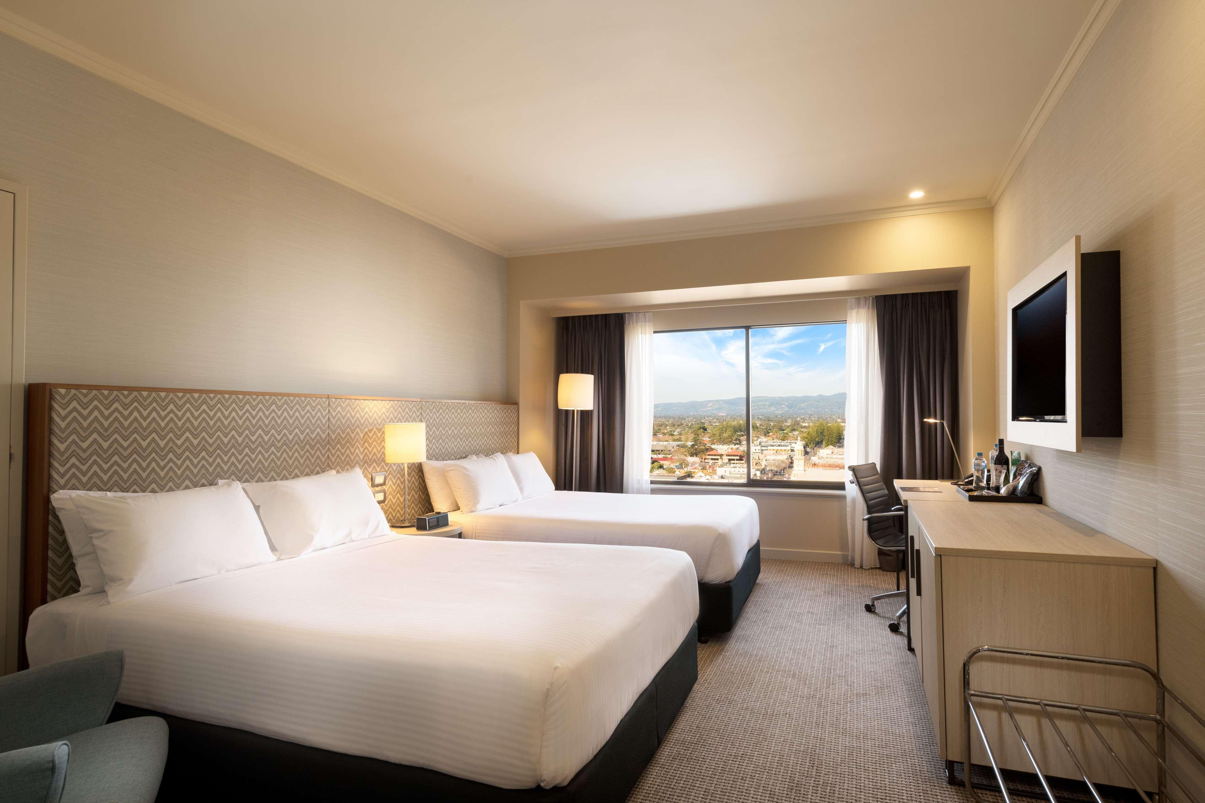 Stamford Grand Adelaide Hotel Гленелг Екстериор снимка The photo shows a modern hotel room featuring two beds with white linens. There are bedside tables with lamps next to each bed. A large window provides natural light and offers a view of the outside, with curtains on either side. The room has a neutr