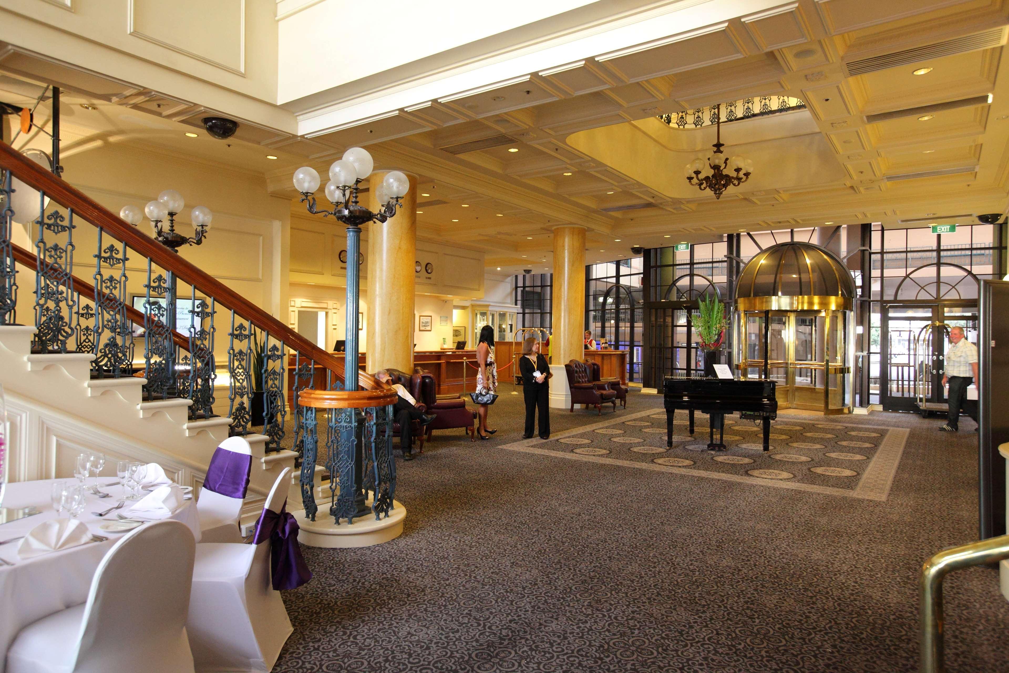 Stamford Grand Adelaide Hotel Гленелг Интериор снимка The photo shows a luxurious interior of a hotel or a restaurant lobby. There is an elegant staircase with ornate railings leading to an upper level. The space features high ceilings with decorative chandeliers and ample natural light coming through l
