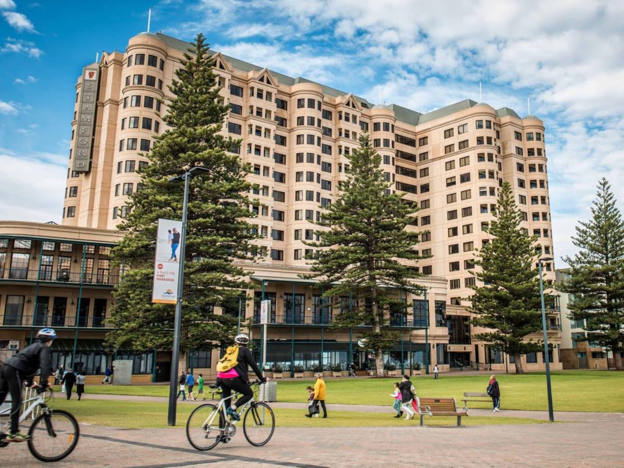 Stamford Grand Adelaide Hotel Гленелг Екстериор снимка The photo depicts a modern building with a large facade, likely a hotel or commercial establishment, situated near a spacious green area. Surrounding the building are several tall trees, providing shade and enhancing the aesthetic appeal. In the fore