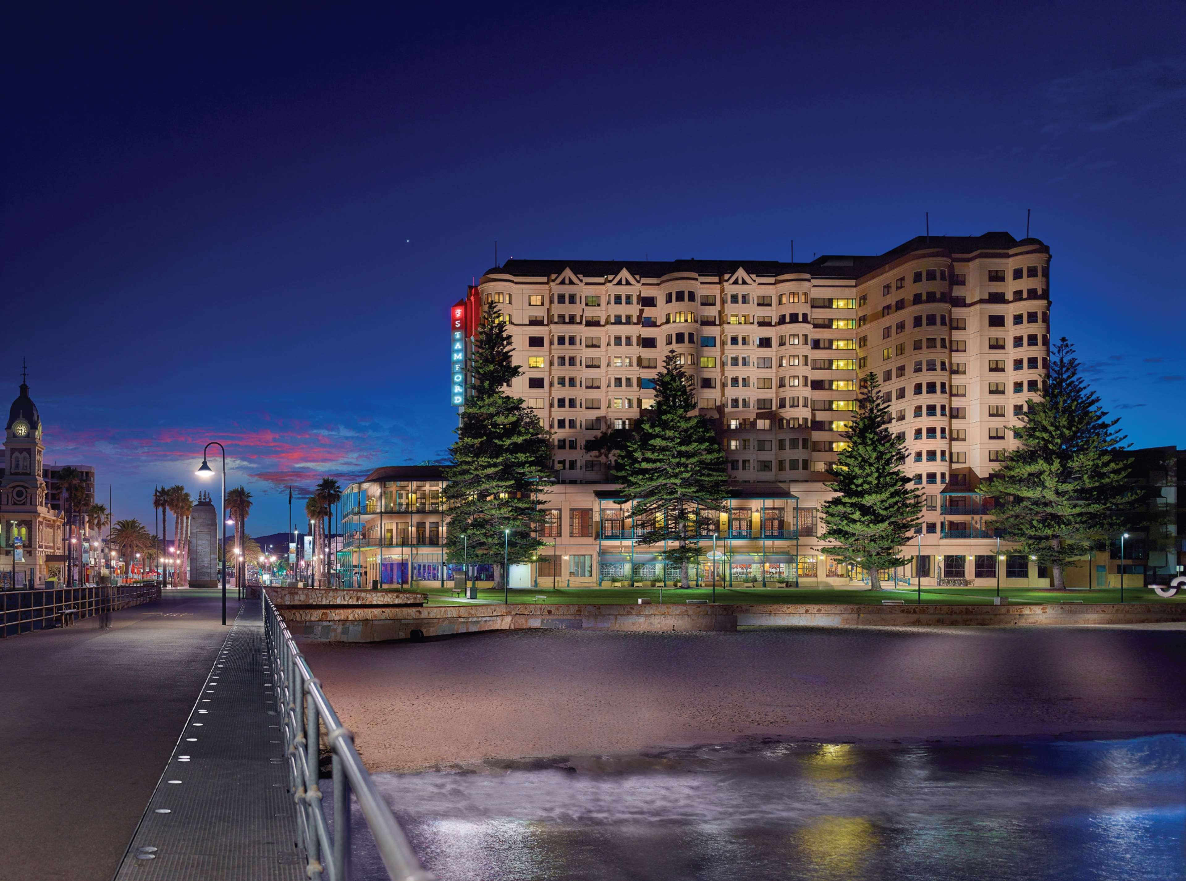 Stamford Grand Adelaide Hotel Гленелг Екстериор снимка The photo shows a waterfront scene at dusk, featuring a large building lit up against a darkening sky. The architecture appears modern and prominent, with several windows illuminated. In the foreground, there is a walkway or pier with fencing along t