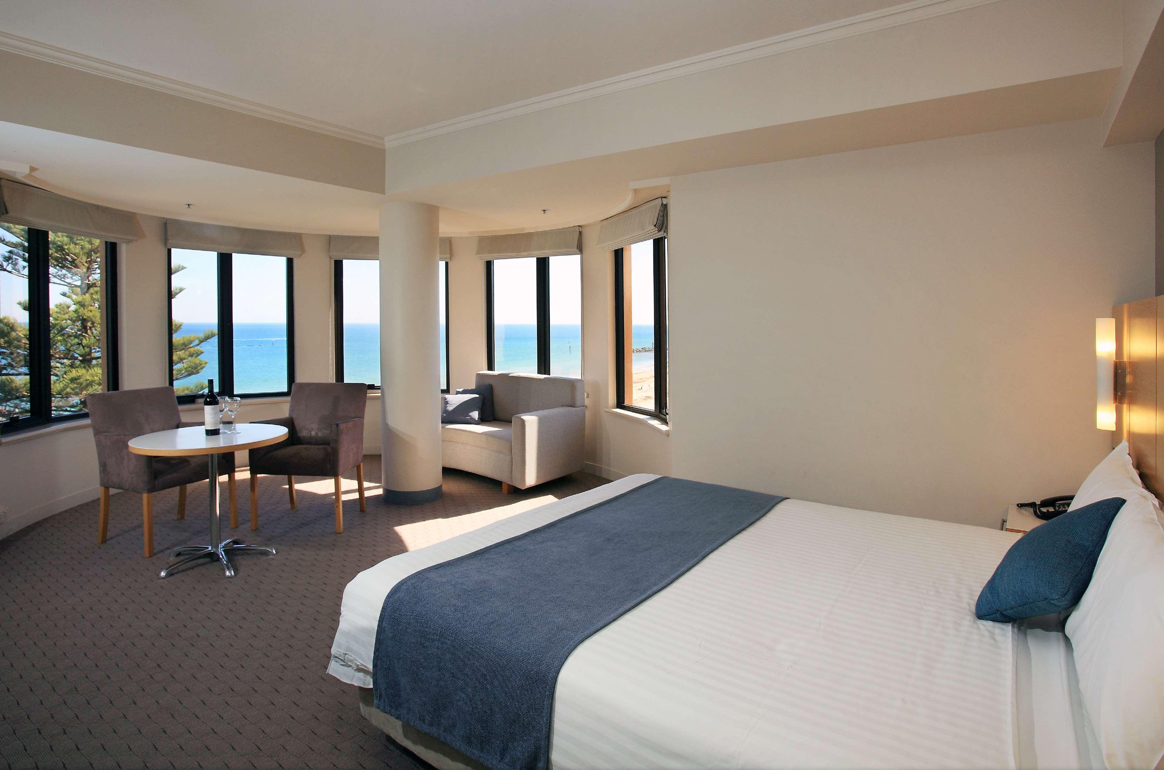 Stamford Grand Adelaide Hotel Гленелг Екстериор снимка The photo shows a well-lit hotel room featuring large windows that offer a view of the ocean. The room is arranged with a comfortable bed covered in white linens and a navy blue throw blanket. There is a small table and chairs set up beside the windo
