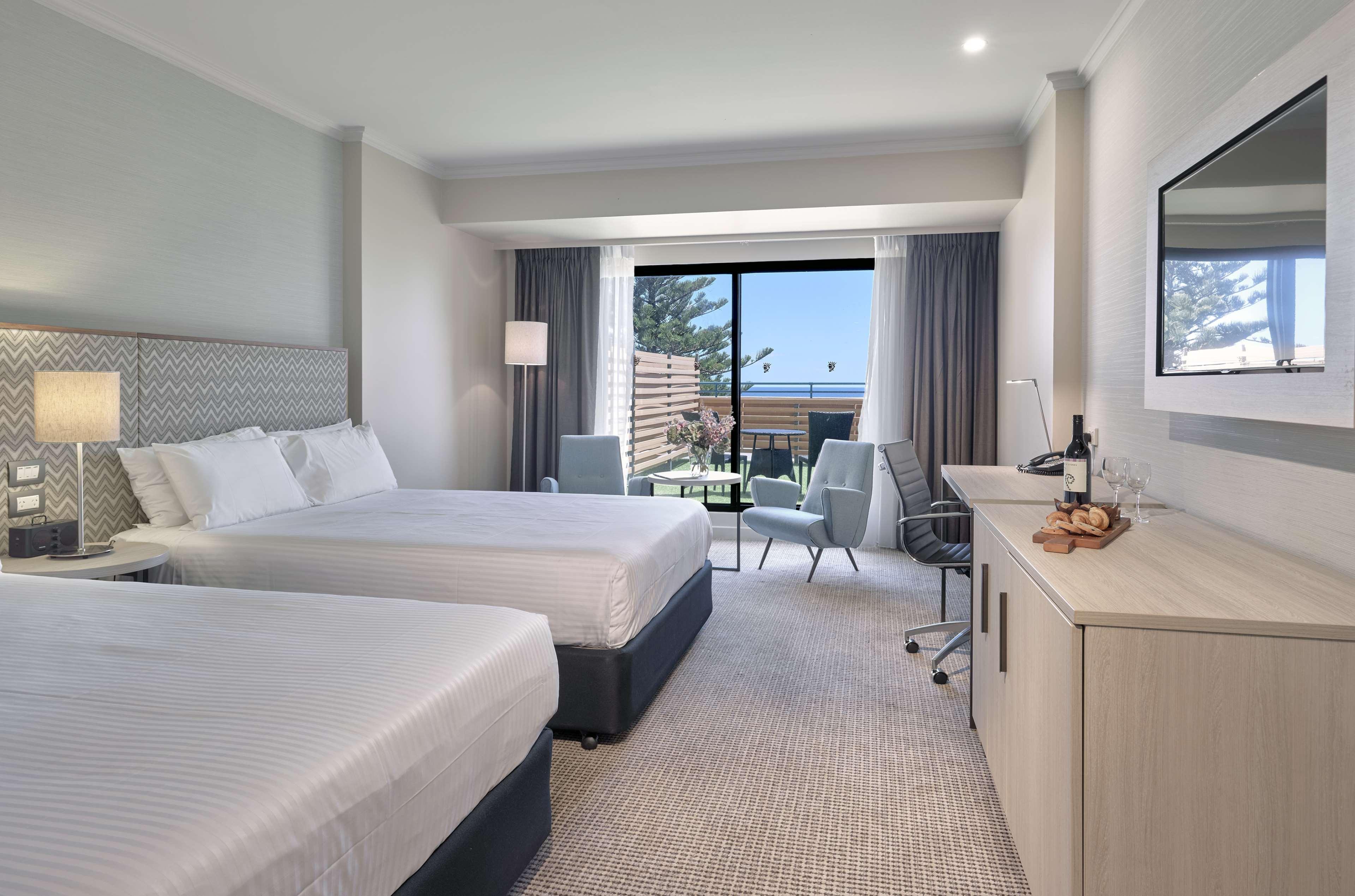 Stamford Grand Adelaide Hotel Гленелг Екстериор снимка The photo shows a modern hotel room featuring two beds with white linens. There's a large window that provides natural light and a view of a balcony area. The room includes two chairs and a small table, creating a cozy sitting area. A television is m