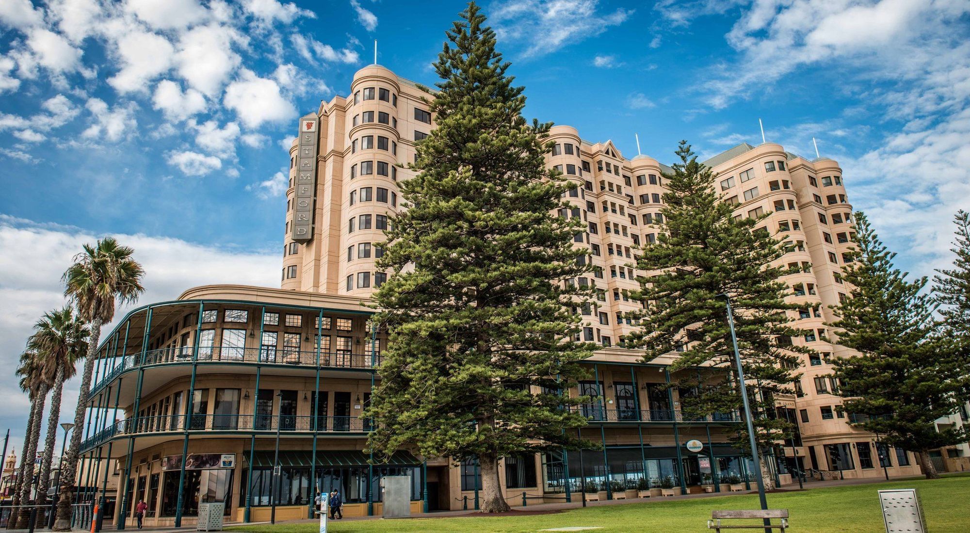 Stamford Grand Adelaide Hotel Гленелг Екстериор снимка The image shows a large building with a distinctive architectural style, characterized by a tan or beige color. The structure appears to have multiple floors and rounded edges. In front of the building, there are several tall green trees, which add t