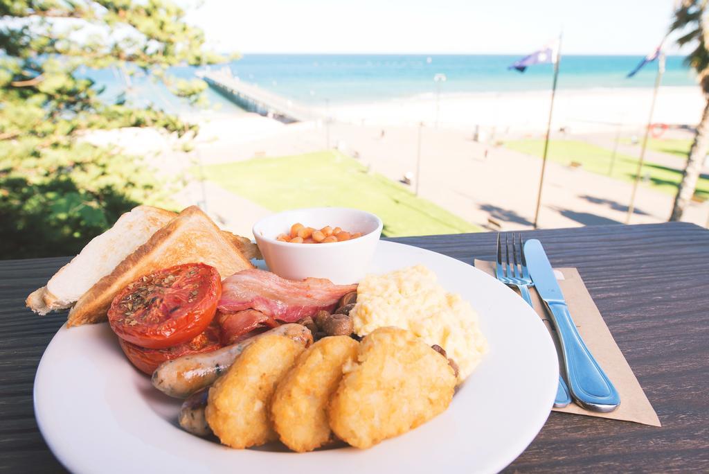 Stamford Grand Adelaide Hotel Гленелг Екстериор снимка The photo shows a plate of breakfast food set against a beachside backdrop. The plate includes several items: crispy bacon, a grilled tomato, sausage links, golden hash browns, and a small serving of baked beans. There is also a piece of toast on the