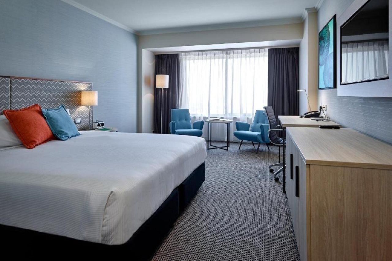 Stamford Grand Adelaide Hotel Гленелг Екстериор снимка The photo shows a modern hotel room with a clean and minimalist design. In the center, there is a large, neatly made bed with white linens and two decorative pillows, one orange and one blue. To the right, there is a small wooden cabinet with a telev